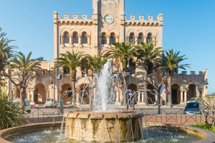 Fountain in a park with town hall building in background in Ciutadella on sunny summer day, Menorca island, Spain